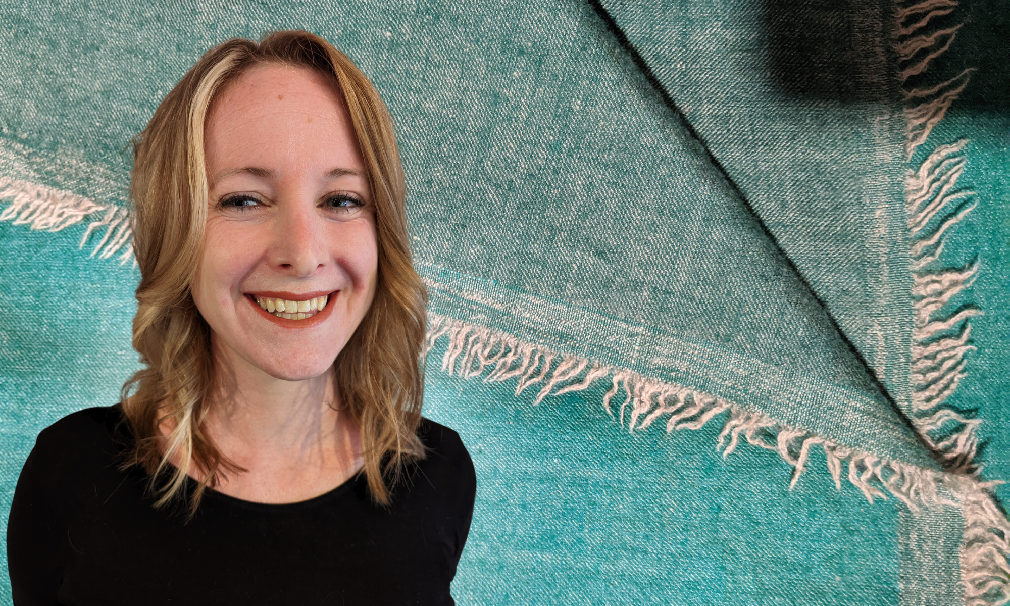 A photo of Heather Elisabeth Lanthorn smiling. She has medium length blonde hair with highlights and is wearing a black shirt. The background is a teal blanket textile.